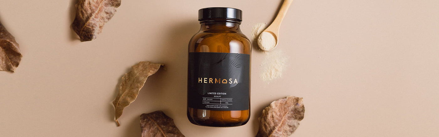 A jar of HERMOSA grass-fed whey protein powder surrounded by leaves.